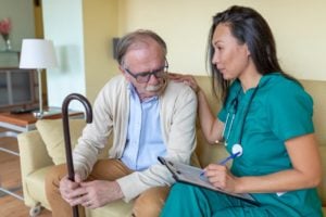 elderly man and visiting nurse with a clipboard sitting on couch talking